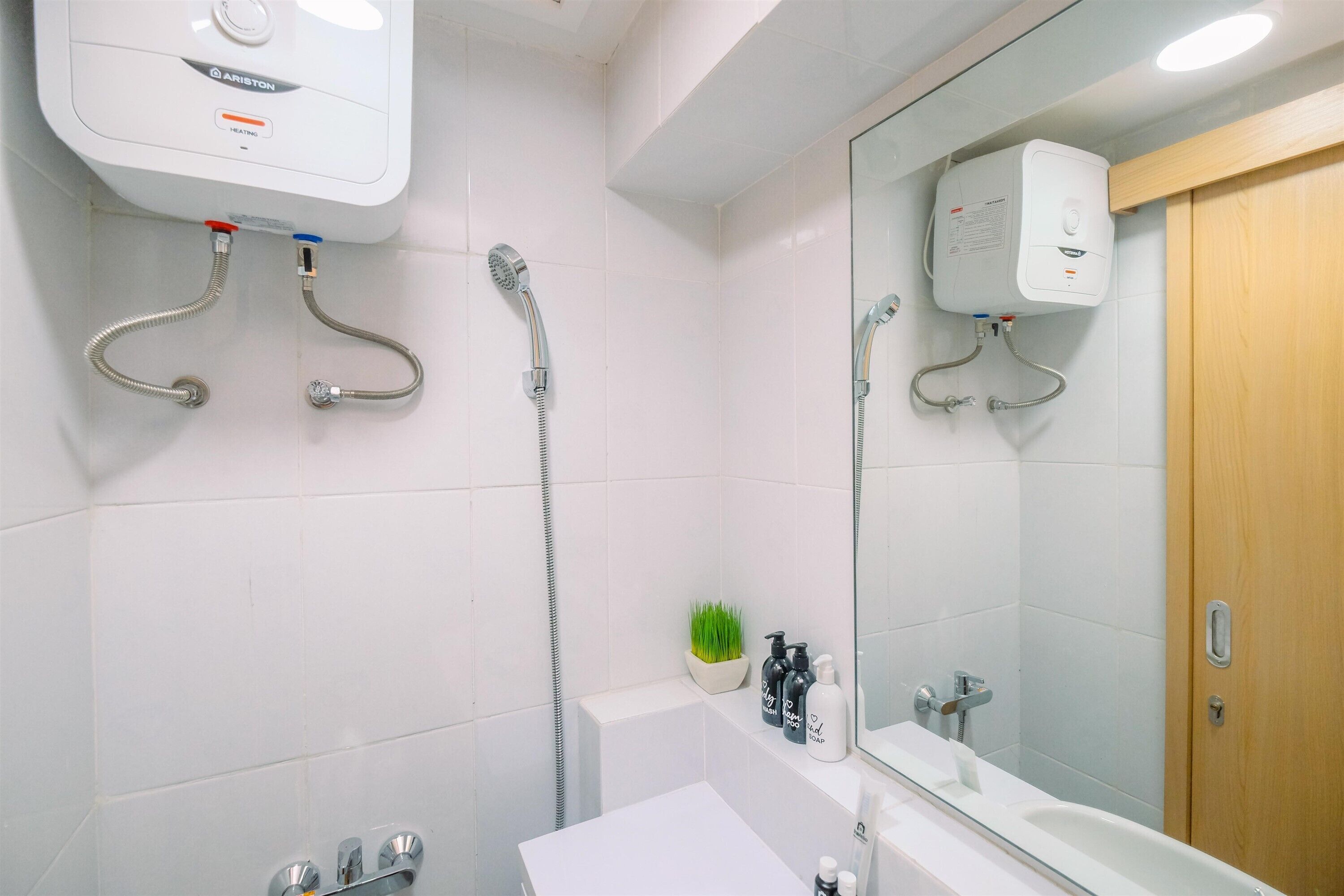 Een moderne badkamer met een witte betegelde muur, een houten deur, een grote spiegel, een wit wastafelmeubel en een douche met een handdouche.
