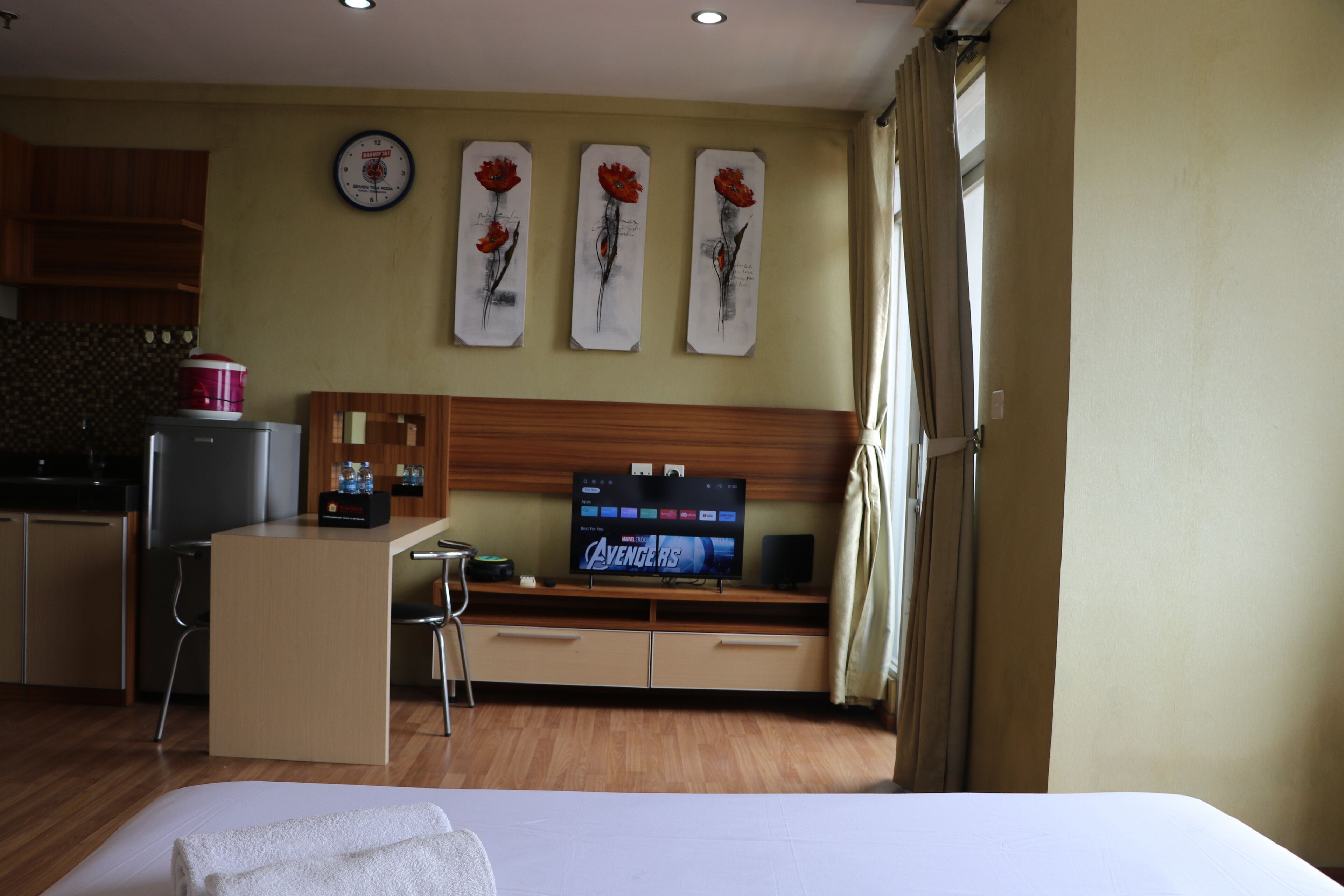 Una camera d'albergo con un letto, una scrivania con televisore che mostra 'Avengers', una sveglia e tre quadri con fiori appesi al muro.