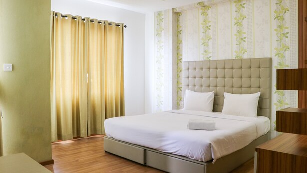 A hotel room with a large bed, a wooden floor, a curtain, and a wall with floral wallpaper. - Spacious Studio The Edge Bandung Apartment (Cimahi)