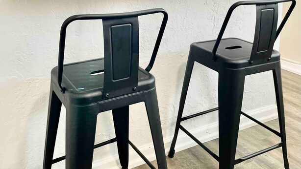 Deux chaises en métal noir avec des pieds en métal, une étagère en bois et une porte blanche dans une pièce aux murs blancs et au plancher en bois.