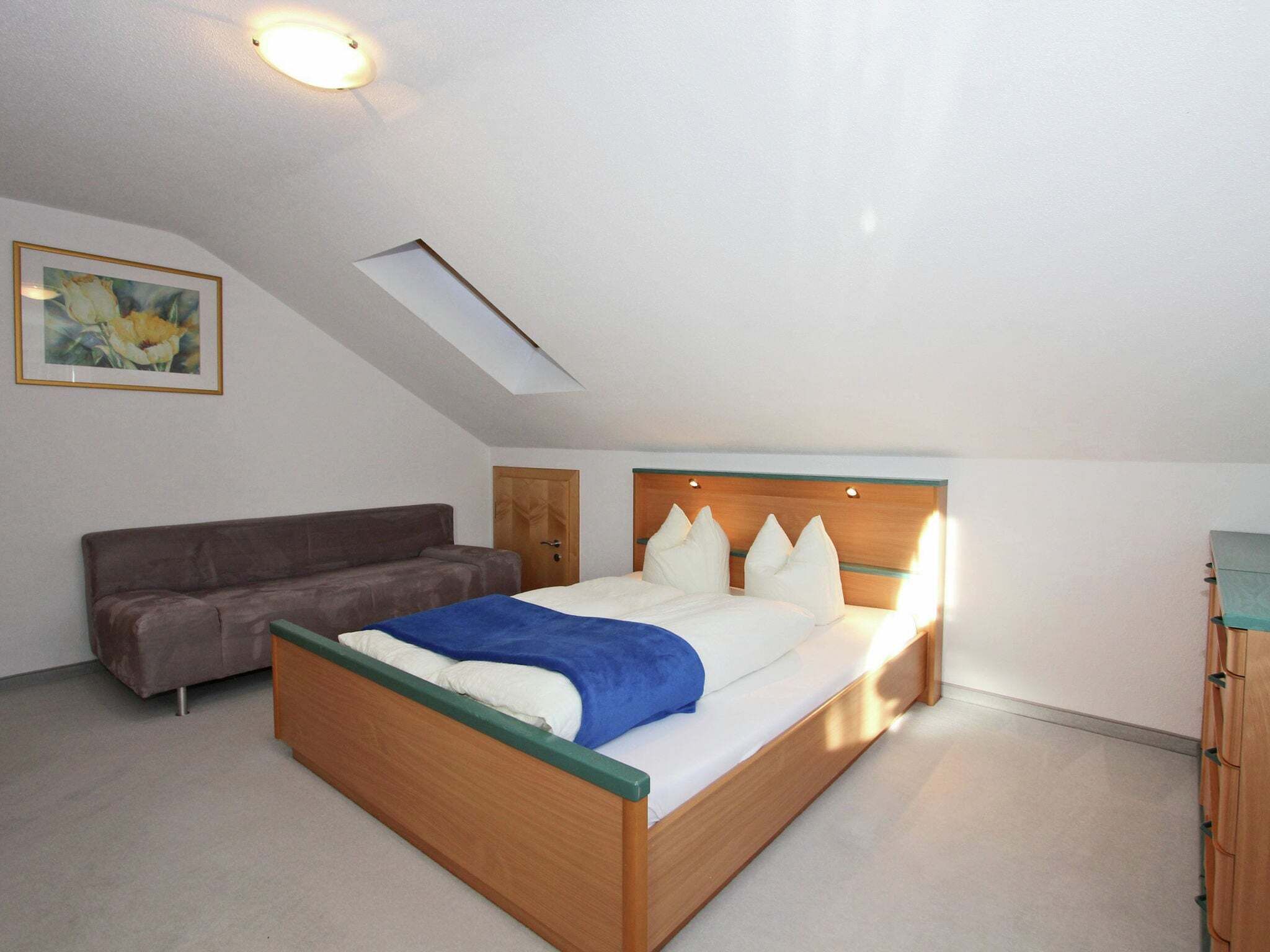 A bedroom with a bed, a sofa, a wooden headboard, a ceiling light, a skylight, and a framed picture on the wall.