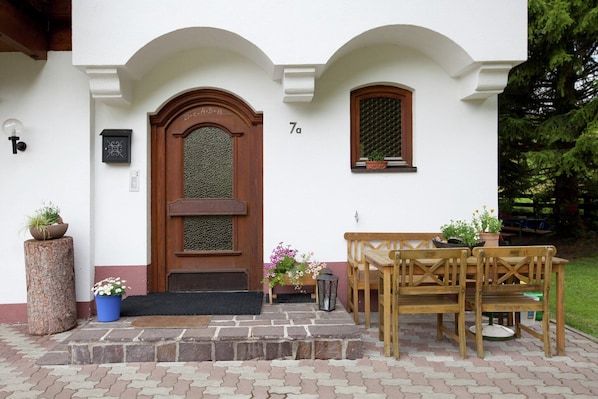 Un edificio blanco con puerta de madera, una banca, una mesa y plantas en macetas.