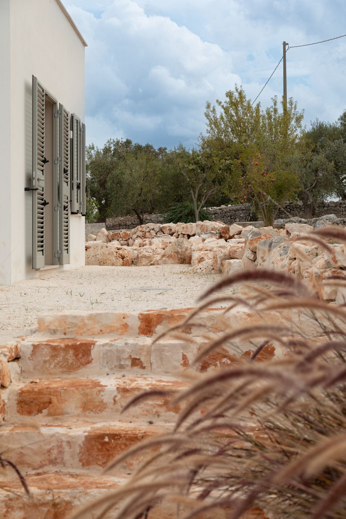 Una escalera de piedra que conduce a un edificio con contraventanas, rodeado de olivos y un muro de piedra.