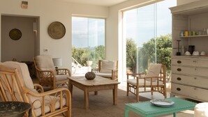 Un salon spacieux, meublé de meubles en osier, avec une table basse en bois et une vue sur la verdure grâce à de grandes fenêtres.