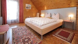 A cozy bedroom with a wooden sloped ceiling, a large bed with floral bedding, two bedside lamps, and a view of the outdoors through a window with sheer curtains. - Apartment in Eberndorf near Petzen Ski Area (Eberndorf)