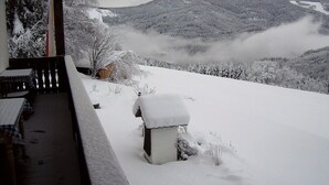 A snow-covered balcony with a wooden bench and a small table, overlooking a snowy landscape with trees and a mountain.