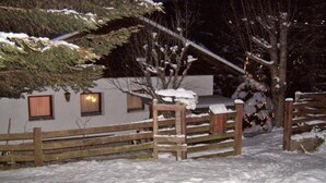 A snow-covered outdoor area with a wooden fence, a snow-covered tree, and a building with windows.