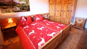 A bedroom with a wooden bed, red bedding with deer patterns, a wooden wardrobe, and a painting of a landscape.