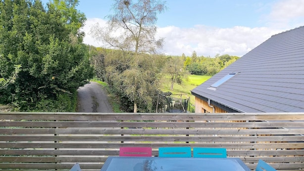 A wooden deck with a table and chairs, overlooking a green landscape with trees and a building with a sloped roof.