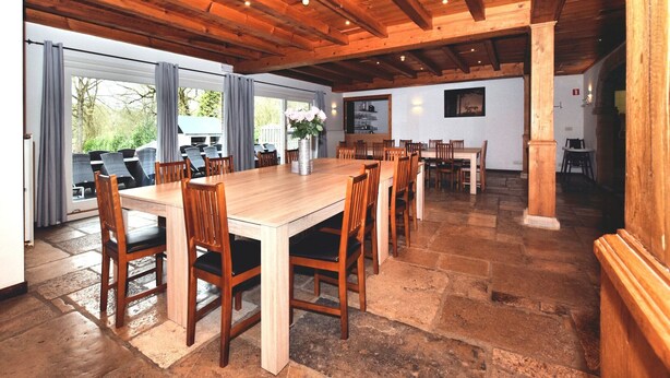 Un vaste espace dédié à la salle à manger, avec une grande table et des chaises en bois, une cuisine à l’arrière-plan et de grandes fenêtres qui laissent entrer la lumière naturelle.