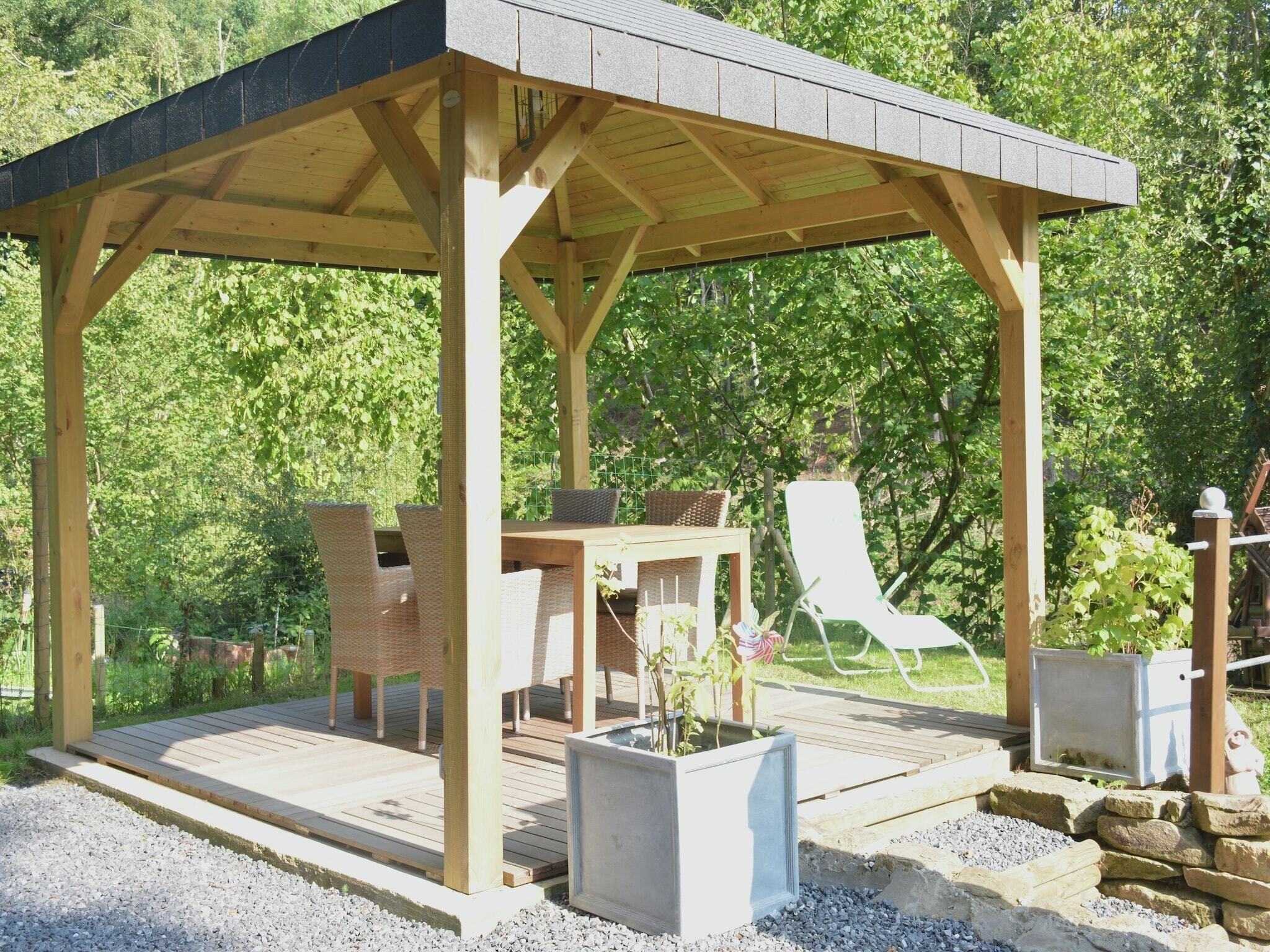 Een houten prieel met een tafel en stoelen, omgeven door grind en planten.