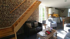 Un espace de vie de style loft avec des murs en briques apparentes, un escalier en bois, un canapé en cuir noir, une table basse en bois et un coin cuisine doté de meubles blancs et d’une décoration représentant une tête de boucher.