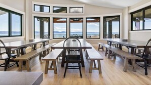 Un comedor con varias mesas y bancos de madera, ventanas amplias con vistas a las montañas y un techo alto con iluminación empotrada.