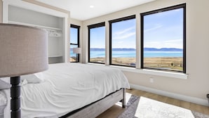 Un dormitorio con una cama grande, una mesita de noche, una lámpara y vistas a un lago a través de amplios ventanales.