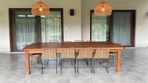 Un vaste espace dédié à la salle à manger, avec une longue table en bois et six chaises en osier, deux ventilateurs de plafond et de grandes portes-fenêtres coulissantes.