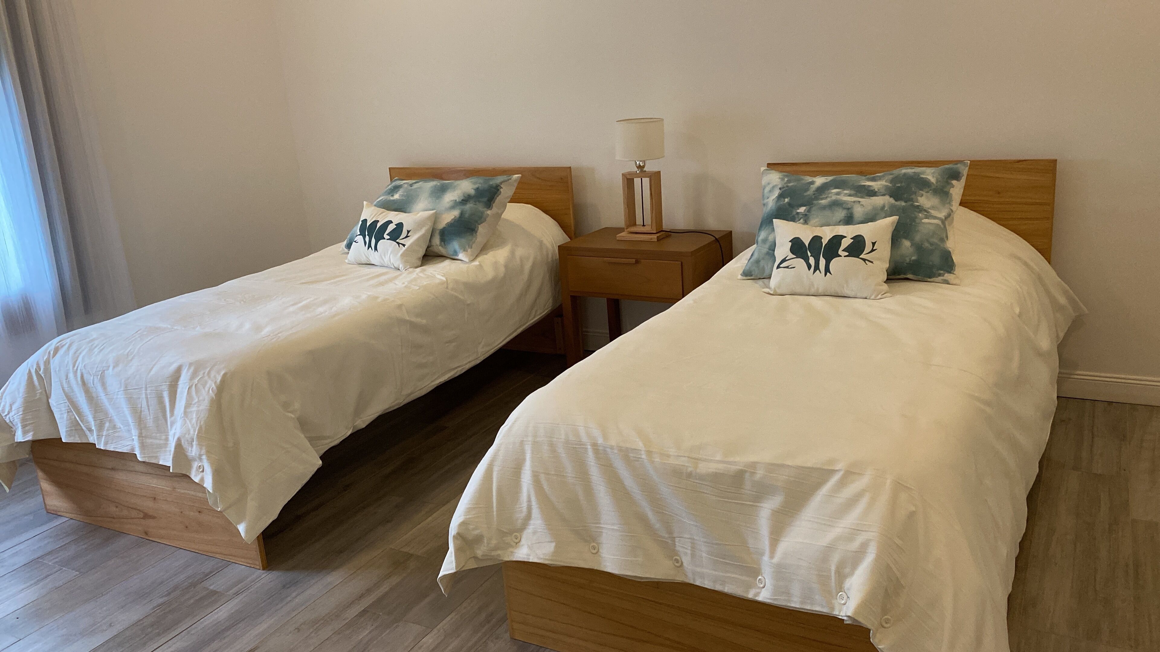 Two single beds with wooden frames and white bedding, each with a pillow featuring a bird design. There is a nightstand with a lamp between the beds.