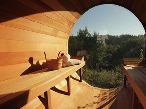 Eine hölzerne Saunabank mit Handtuch und Holzschale, vor einer Baumlandschaft und einem entfernten Gebäude.