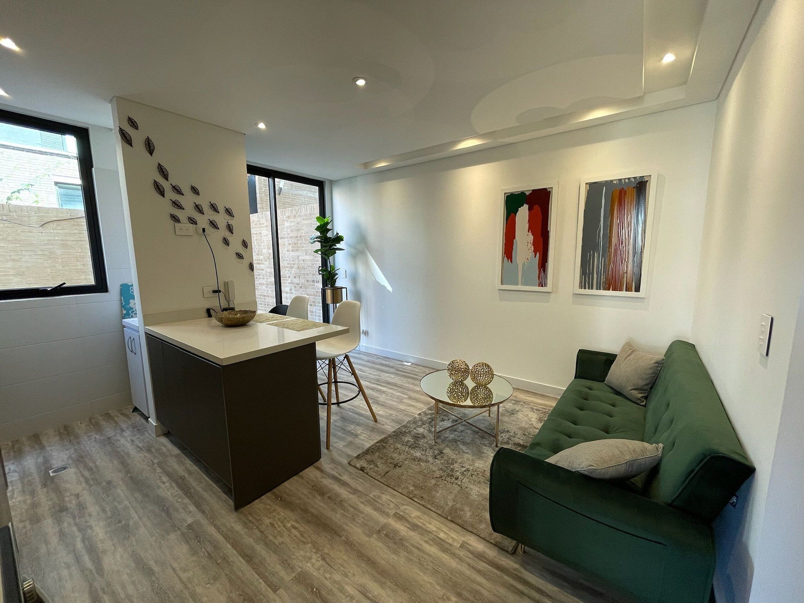 Een moderne woonkamer met een groene bank, een klein rond salontafeltje, en een keukenhoek met een wit aanrecht en een spoelbak.