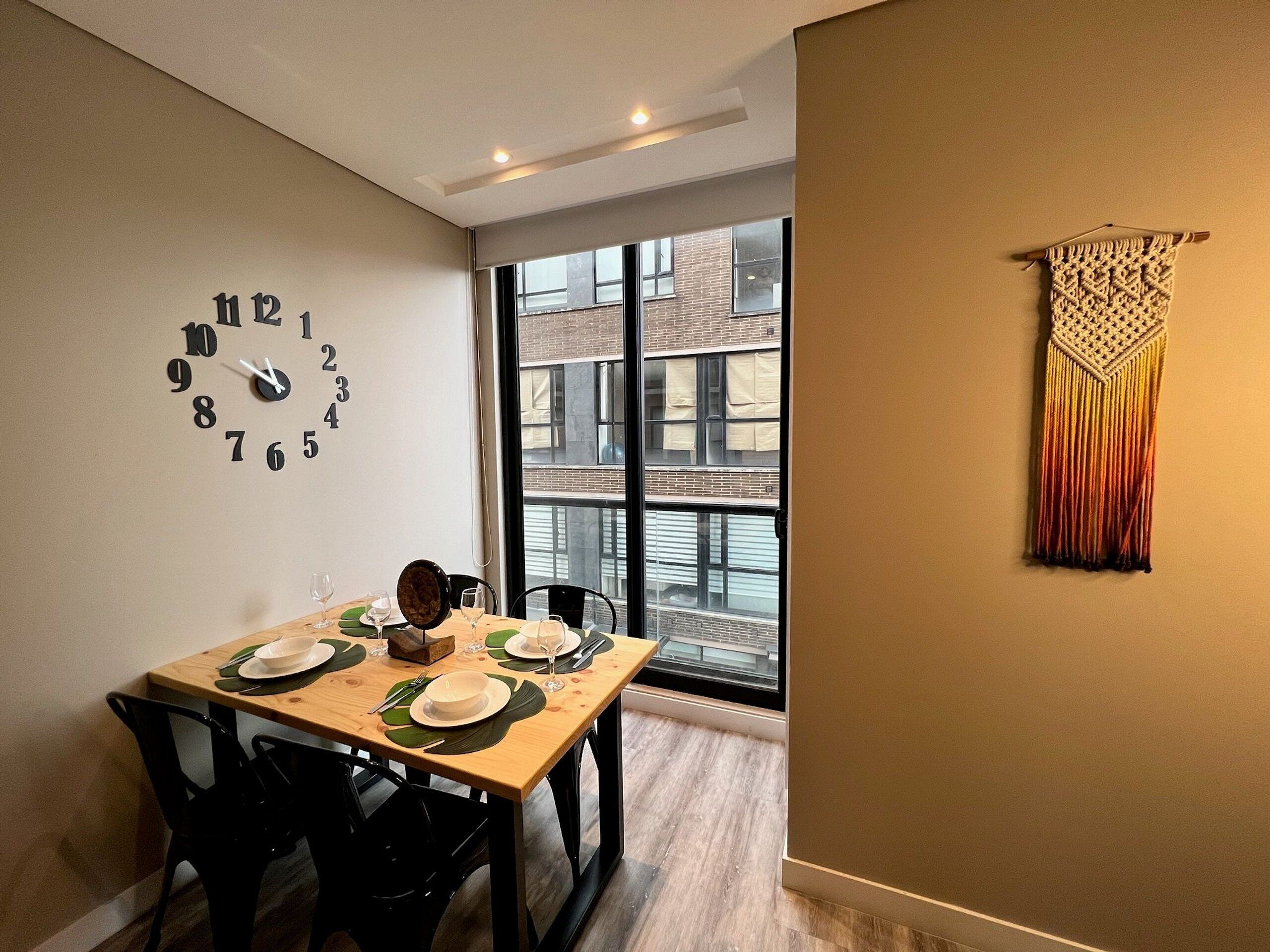 Un coin repas avec une table en bois dressée pour quatre personnes, une horloge murale et une fenêtre donnant sur des immeubles.
