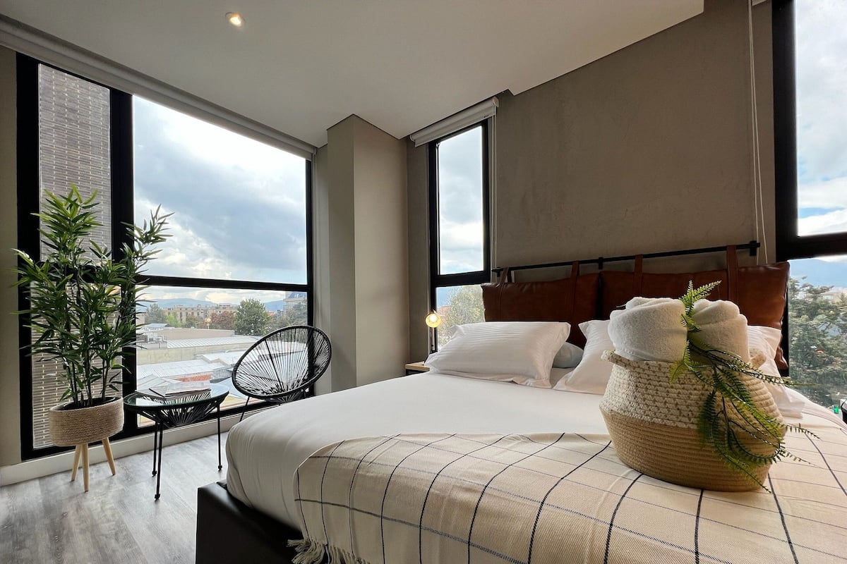 A modern bedroom with a large bed, a view of the city through large windows, a potted plant, and a chair.