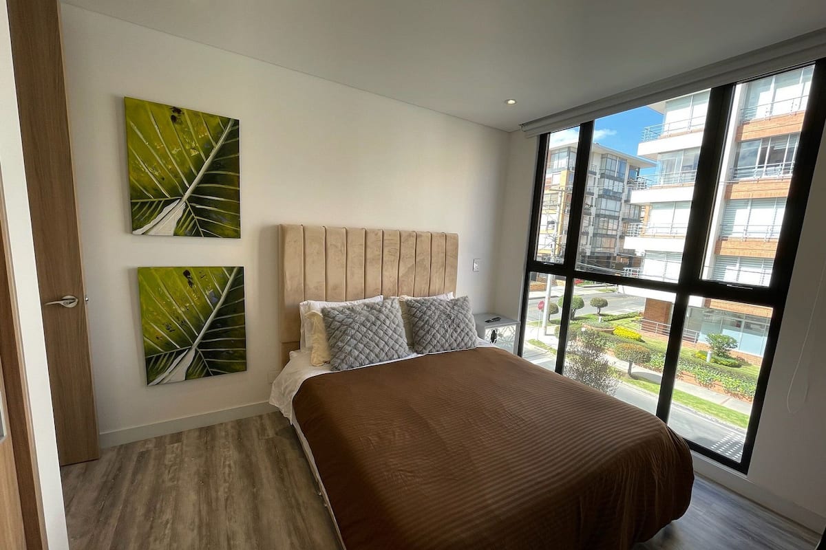 A modern bedroom with a large bed, two wall-mounted artworks, and a view of the city through large windows.