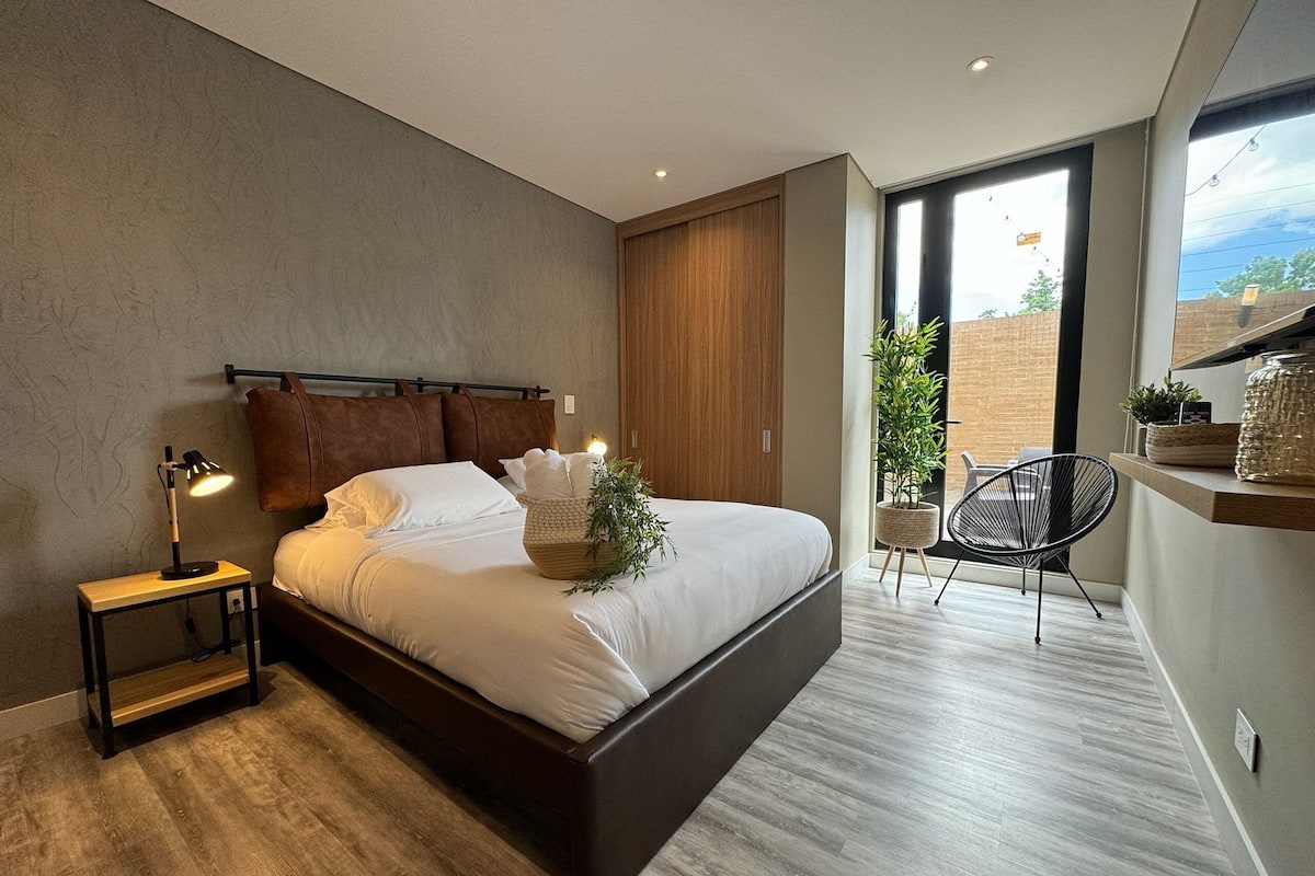 A modern bedroom with a large bed, a bedside table with a lamp, a chair, and a view of the outdoors.