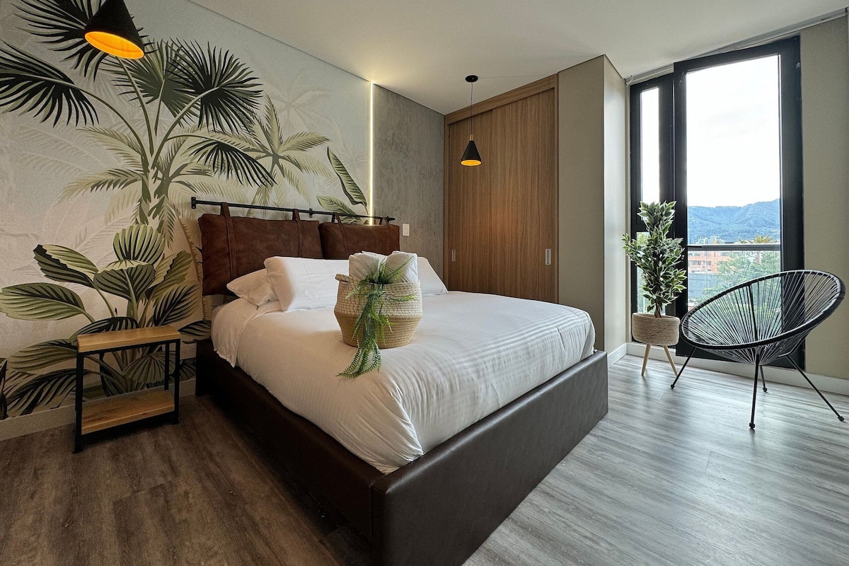 A modern bedroom with a large bed, a wooden headboard, a nightstand, a black chair, a potted plant, and a view of the outdoors through a large window.