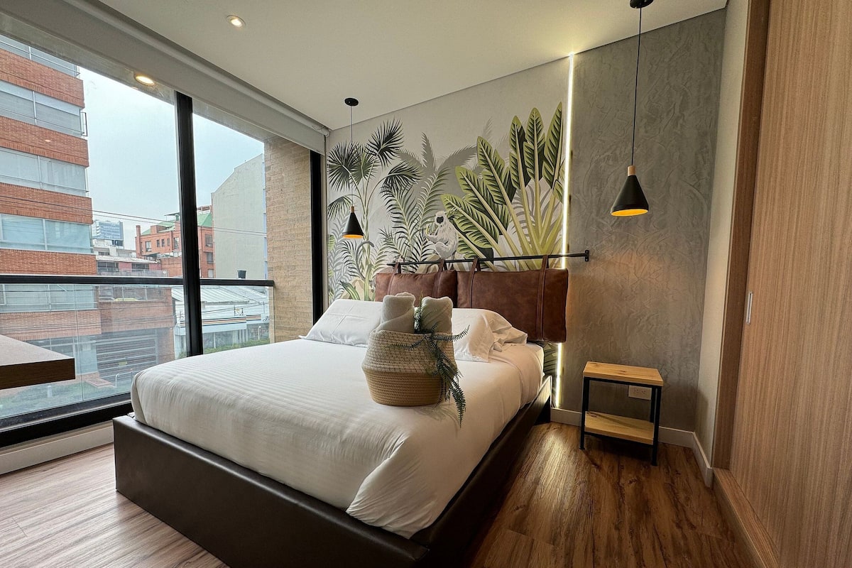 A modern bedroom with a large bed, a view of buildings through a window, and a plant wall.
