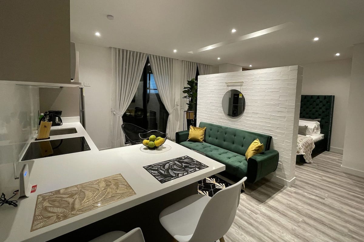 A modern living room with a green sofa, a dining table with a fruit bowl, and a white wall with a textured pattern.