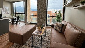 A modern living room with a sofa, ottoman, and coffee table, large windows with a city view, and a kitchen area.