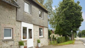 A two-story stone house with a white door and windows, a small garden with potted plants, and a large tree in front. - Holiday Home with Garden, Heating, Barbecue (Butgenbach)