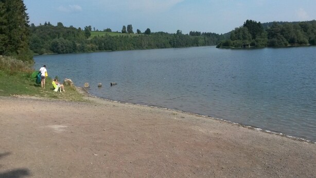 A lakeside view with a dirt path leading to a grassy area where people are sitting. - Holiday Home with Garden, Heating, Barbecue (Butgenbach)