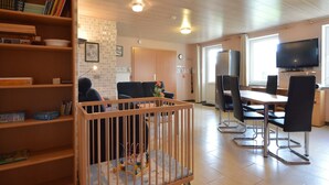 Un salon avec un berceau en bois, un coin repas avec une table et des chaises, et un téléviseur fixé au mur.