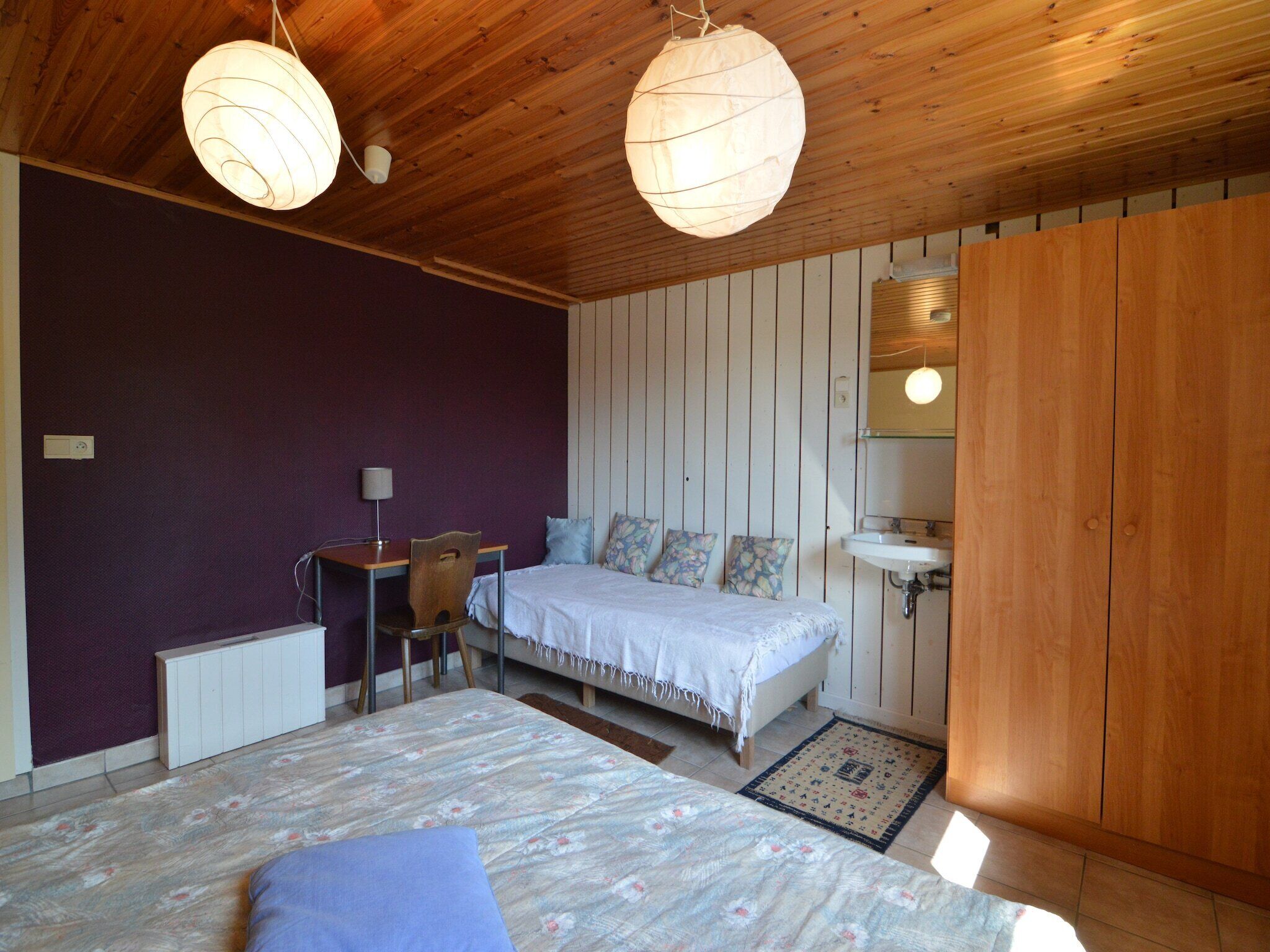 A bedroom with a wooden ceiling, a bed with floral bedding, a desk with a chair, a bedside table, and a mirror.