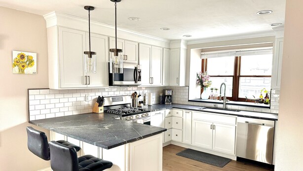 A modern kitchen with white cabinets, a black countertop, stainless steel appliances, and a bar stool. - Victorian Village Townhome, large living space in the heart of the short north! (Columbus)