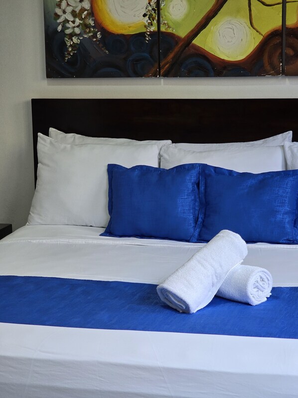 A neatly made bed with white and blue bedding, a rolled white towel, and a painting above it. - Tropical Paradise (Jacó)