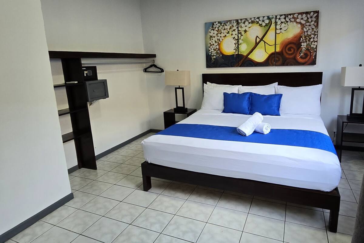 A hotel room with a bed, a painting on the wall, and a desk with a lamp.