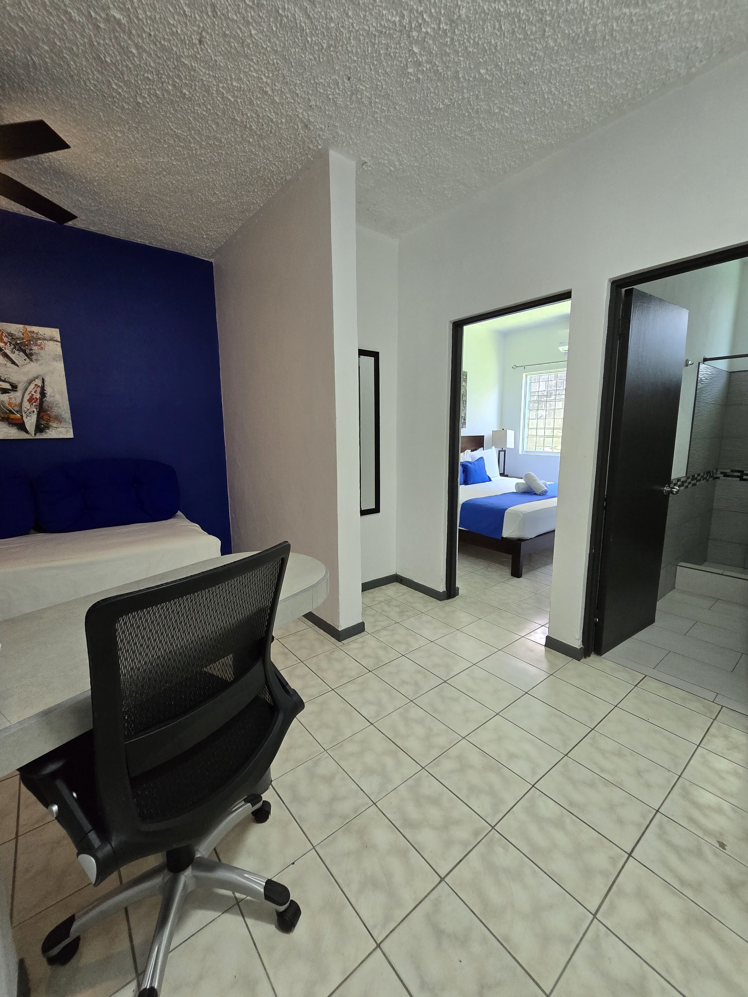 A hotel room with a bed, a desk, and a chair. There is a bathroom with a shower and a toilet. The room has white walls and a blue accent wall.