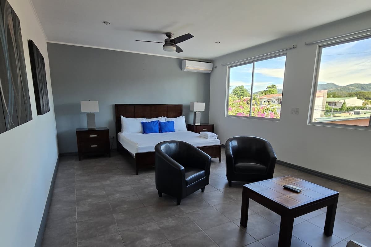 A hotel room with a bed, two armchairs, a small table, a ceiling fan, and large windows offering a view of the outdoors.