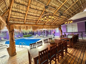 An indoor pool area with a thatched roof, wooden tables and chairs, and a view of the pool. - Tropical Paradise (Jacó)