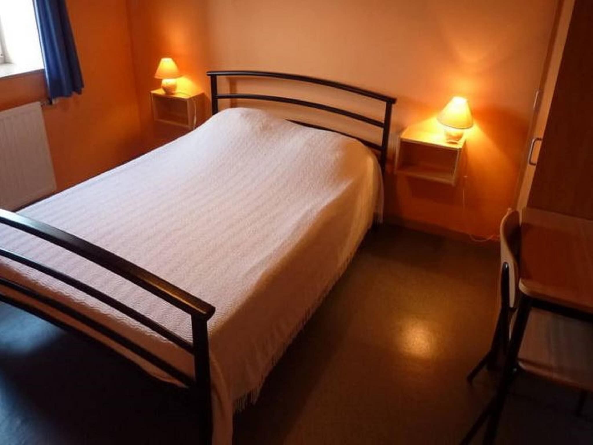 Een eenpersoonsbed met houten frame, twee nachtkastjes met lampen en een stoel in een kamer met oranje wanden en een radiator.