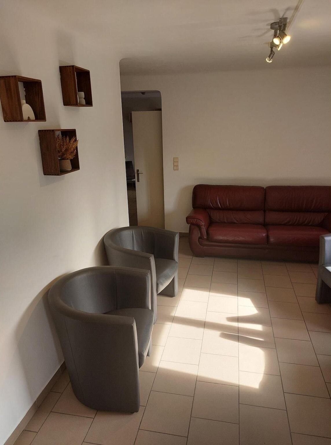 Una sala de estar con un sofá de cuero marrón, dos sillones grises y estanterías de madera en la pared.