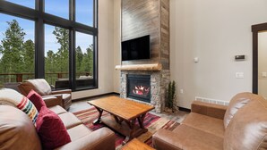 A spacious living room with leather sofas, a wooden coffee table, a stone fireplace, and large windows offering a view of trees. - Stylish home with hot tub, pool access, wifi and garage (Lead)
