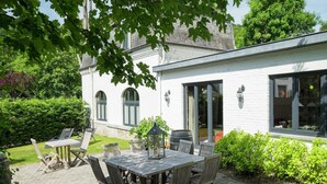 A patio with a wooden table and chairs, surrounded by greenery and a white building with arched windows. - Namur Mansion by the Meus (Hastière par Dela)