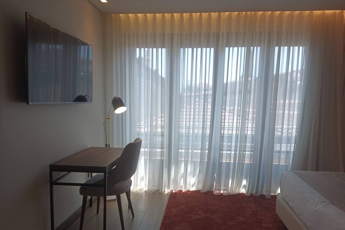 A hotel room with a bed, a desk with a chair, a TV mounted on the wall, and sheer curtains covering the window.
