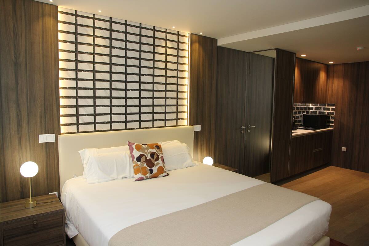 A modern bedroom with a large bed, a nightstand, a wall-mounted light, and a built-in kitchen area.