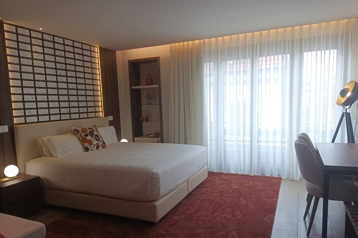 A modern hotel room with a large bed, a desk with a chair, a bookshelf, and a large window with sheer curtains.
