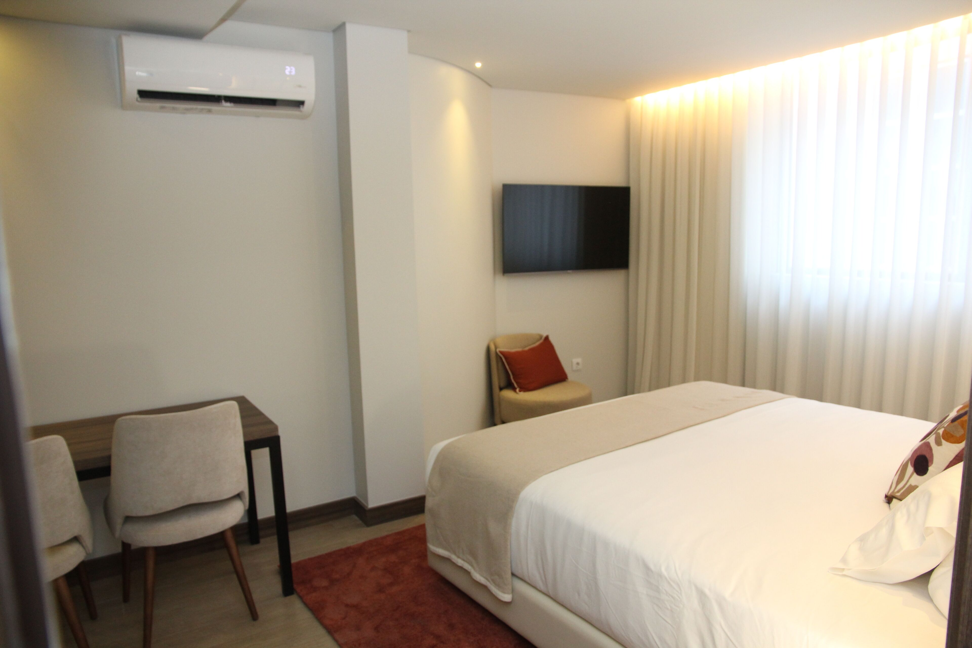 Habitación de hotel con cama, escritorio, silla, televisor montado en la pared y ventana con cortinas.