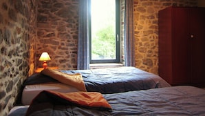 A bedroom with stone walls, a wooden ceiling, a bed with blue bedding, a wooden wardrobe, and a window with curtains.