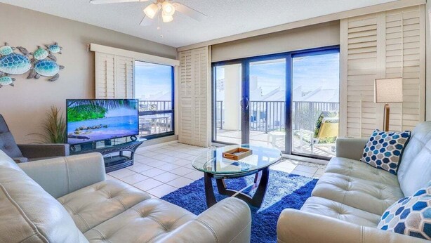 A living room with a television, leather sofas, a glass coffee table, and a balcony with a view. - Silver Sands Sanctuary: Ocean + Pool Balcony Views (New Smyrna Beach)